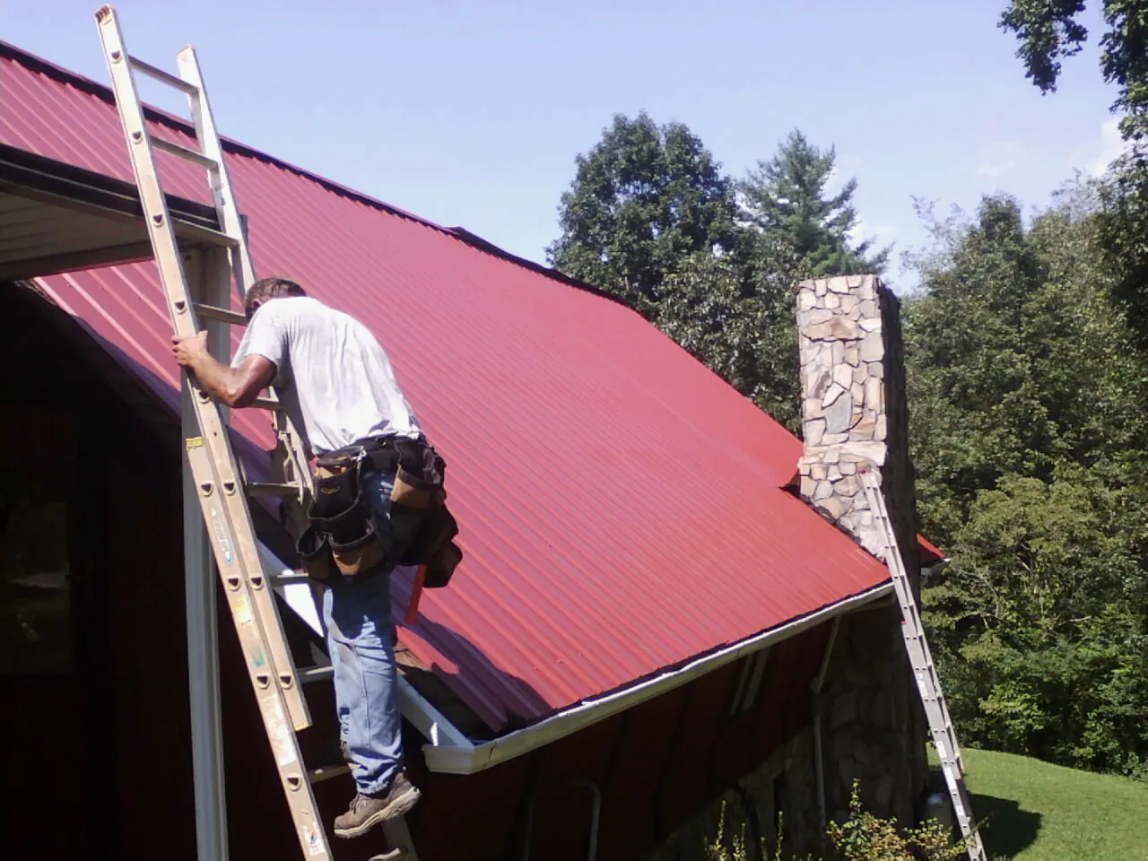 Roofer on a ladder beside a red metal roof and stone chimney — Soffit & Fascia Repair by Roofing Brothers in La Grange Park
